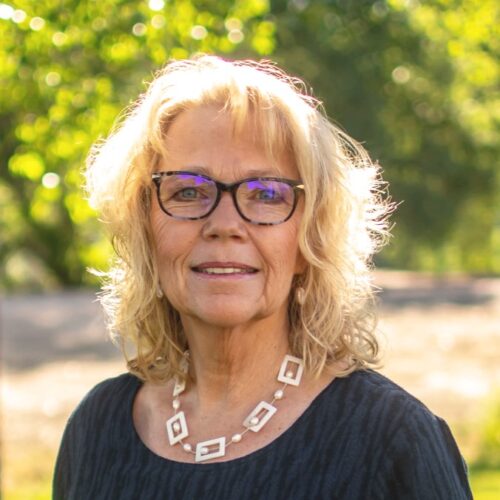 Blonde woman with glasses in front ofgreen foliage