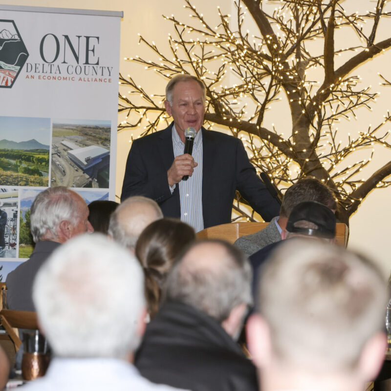 Greg Pope, speaks tot eh audience next to One Delta Countys banner and a tree filled with twinkle lights.